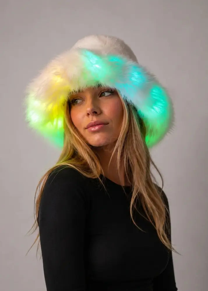 A woman wearing a fluffy white hat with glowing LED lights in green, yellow, and blue, set against a plain gray studio background.