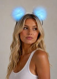 A woman with long blonde hair wearing a white ribbed tank top and glowing blue fluffy LED pom-pom headband against a neutral beige background.
