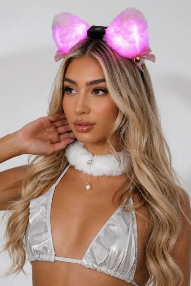 A woman wearing a silver metallic bikini top, a white faux-fur collar necklace with a pearl pendant, and glowing pink bunny-ear headband against a light gray studio background.