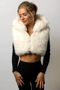 A smiling model wearing a cropped, white faux fur vest with a large hood. She is holding the edges of the hood near her face and wearing a black long-sleeve top underneath and black yoga pants.