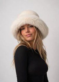 A woman wearing a fluffy cream-colored fur hat and a black long-sleeve top, photographed against a plain light gray studio background.