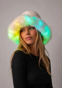 A woman wearing a fluffy white hat with glowing LED lights in green, yellow, and blue, set against a plain gray studio background.