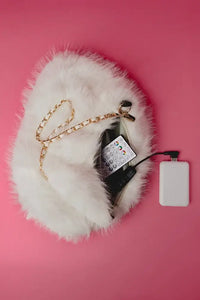 A fluffy white faux-fur handbag with a gold chain strap, shown from above on a pink background with a remote control and a white power bank placed inside and beside it.